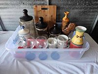 Plastic bin showing the assorted kitchen jars, glassware, mugs, wooden cutting board and ceramic pitchers