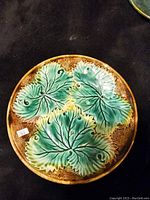 Top view of circular Majolica plate showing three large green leaves on brown speckled background with embossed texture and veining details.