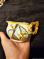 Photo of the front side of the antique creamer showing the hummingbird, fan, and flower relief decoration and ornate handle.