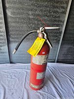 Full view of red metal fire extinguisher showing hose attached to the neck and handle with attached yellow inspection tag.