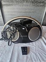 Front view of black Harman/Kardon radio with silver speaker grilles and handle, power adapter resting on top, and remote control placed in front.