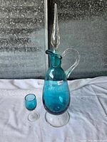 Blue glass decanter with clear glass handle, matching glass cup, both on white fabric, black background