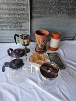 Overview of all items showing three coffee pots, thermos, ceramic vase, leaf dish, and grater on white cloth.