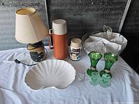 Wide angle photo of all items including ceramic lamp, orange insulated container, Six Million Dollar Man thermos, green glass cruet set, white scallop dish and glassware.