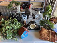 Wide view of lot with decorative black mantle clock, plants, white bird cages, small painted birdhouse, and fabric cushions on display surface