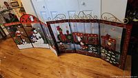 Four connected metal fireplace screens with painted Christmas scenes including snowmen, train, fireplace, and holiday ornaments, displayed indoors on a wooden floor.
