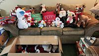 Full view of sofa with multiple Christmas-themed plush toys, throw pillows, blankets, and towels arranged, showing quantity and variety of items.