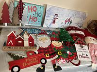 Image shows a tabletop display of varied Christmas decorations including wooden trees, Santa and elf cutouts, festive signs, and holiday greenery.