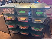 Full view of the metal rack holding 12 plastic storage bins with green lids, each bin labeled with contents such as Pom Poms, Glitter, Feathers, Dice Counter Chips, and others.