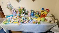 Wide view of a table covered in a light blue cloth holding multiple Easter decor items including plastic eggs, bunny figurines, wreaths, trays, gel clings and seasonal decorations