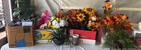 Photo showing a collection of faux floral arrangements in fall colors along with other faux flowers and greenery, in various containers and boxes.