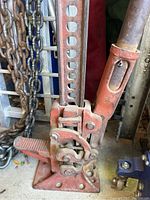 Red mechanical farm/off-road vehicle jack with a base plate, lever, and vertical adjustable steel beam with holes for height adjustment, alongside a metal chain accessory.