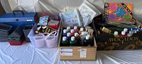 Wide view of assorted kids arts and crafts supplies including bottles of paint, paintbrushes, plastic containers, and closed boxes of craft kits.