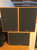 Three Klipsch speakers front view showing oak-oil veneer and black grilles: two satellites and one center channel.