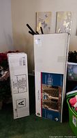 Two boxed artificial Christmas trees, one taller and one shorter, standing upright with packaging showing images of green pre-lit trees.