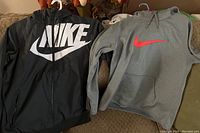 Two Nike outerwear items hanging side by side; black and gray windbreaker jacket with a large white Nike logo and gray pullover hoodie with red Nike swoosh logo.