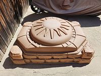 Top and angled side view of small plastic sandbox with sun design on lid.