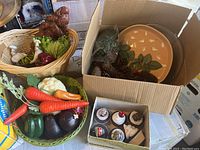 Photo showing two baskets with various fake vegetables and a small box of decorated fake cupcakes along with a cardboard box containing a ceramic plate and pine cones.