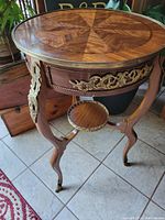 Side view of table showcasing three curved legs with gold metal appliques, bottom tier shelf, and sunburst patterned veneer top with braided trim edge.