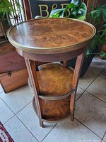 Full view of the round 3-tier antique wood side table showing veneer tops and brass edging, carved leaf leg detail.