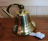 Side profile of a polished brass ships bell with attached brass mount bracket and knotted white rope pull lying on a wooden surface.
