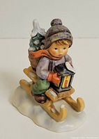 Front view showing child in winter clothes holding lantern on sled with basket of snow-covered trees and bottles.