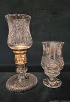 Pair of clear crystal hurricane lamps, one large with brass components and one smaller all-crystal, shown side by side on a dark surface.