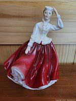 Front and side view showing the figure in glossy white jacket and red pleated skirt, hand lifting small hat.