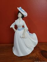 Front and partial side view of Royal Doulton bone china figurine 'Margaret' featuring a woman in white dress with blue sash and hat.