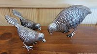 Three silver cast metal bird figurines on a wooden surface against a wood panel background, showing two smaller birds with raised tails and a larger bird with a rounded body.