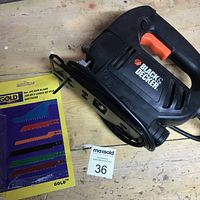 Black & Decker jigsaw and new pack of assorted blades laid on wood surface, showing side profile of the jigsaw and blade pack