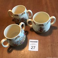 Three Royal Doulton Bunnykins two-handled bone china mugs arranged on a wooden surface, showing front and top views of the mugs with colorful Bunnykins illustrations.