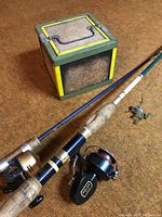 Photo showing two fishing rods with cork handles and reels alongside a vintage Oberlin metal bait canteen box with green and yellow trim and cork panels