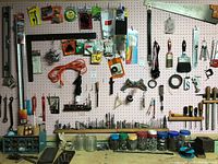 Wide view of pegboard with assorted tools, brushes, files, paint scrapers, saw, and jars of fasteners below on bench