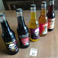Five vintage collectible soda pop bottles with unique skull and graphic labels, sealed and unopened, standing on wooden table.