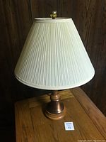 Table lamp with wood and copper base and pleated cream fabric shade, standing on wooden surface against wood panel wall.