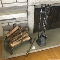 Wrought iron fireplace tool set standing beside a metal wood tote basket filled with cut firewood, set on hearth in front of brick fireplace.