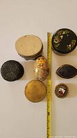 Seven assorted small trinket boxes arranged on flat surface with measuring tape to indicate sizes