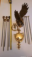 Photo of all lot items including brass eagle, door knocker, horse brass, and skewers alongside measuring tape showing scale.