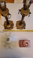 Top-down view showing pair of brass candlesticks, white glass lampshade, and pink vintage alarm clock, with measuring tape showing approximately 13 inch height of candlesticks