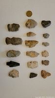 Photo showing a collection of 17 North American flint and stone arrowheads in various shapes and sizes, with a coin for scale.