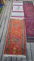 Three rugs displayed on wooden deck: Mexican style rug with striped pattern at top, orange/red geometric floral runner at left, dark red Persian style runner at right