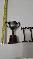 Silver trophy cup with wooden base alongside a pair of unusual silver cutter rests, measuring tape shows trophy height approximately 18 cm.