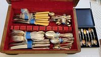 Top view of mixed antique silverware organized in an old red felt lined wooden cutlery box showing various knives, forks, spoons and other utensils wrapped in blue tape and protective tissue.