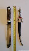 Two cutters displayed with a tape measure for size reference; one large Bowie-style with stag-style handle, one Middle-Eastern style curved blade with ornate silver-tone handle and black sheath with decorative tip.