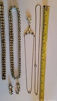 Five pieces of vintage jewelry including a rhinestone bracelet, rhinestone necklace, pendant necklace with chain, additional chain, and pair of stud earrings, all in white metal with clear glass stones. Measuring tape shown for scale.