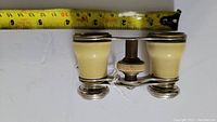 Top-down view of vintage opera glasses showing cream-colored barrels and silver accents with measuring tape indicating width just over 3 inches.