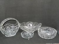 Four clear cut crystal or pressed glass pieces on a black background: a handled basket, oval bowl, round bowl, and small dish.