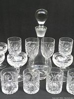 Front-facing image of one large crystal decanter with a stopper surrounded by clear cut crystal glasses in the lot.