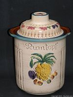 Full view of vintage Rumtopf jar with lid, showing cream-colored stoneware body with raised lettering and hand-painted fruit motif including pineapple.
